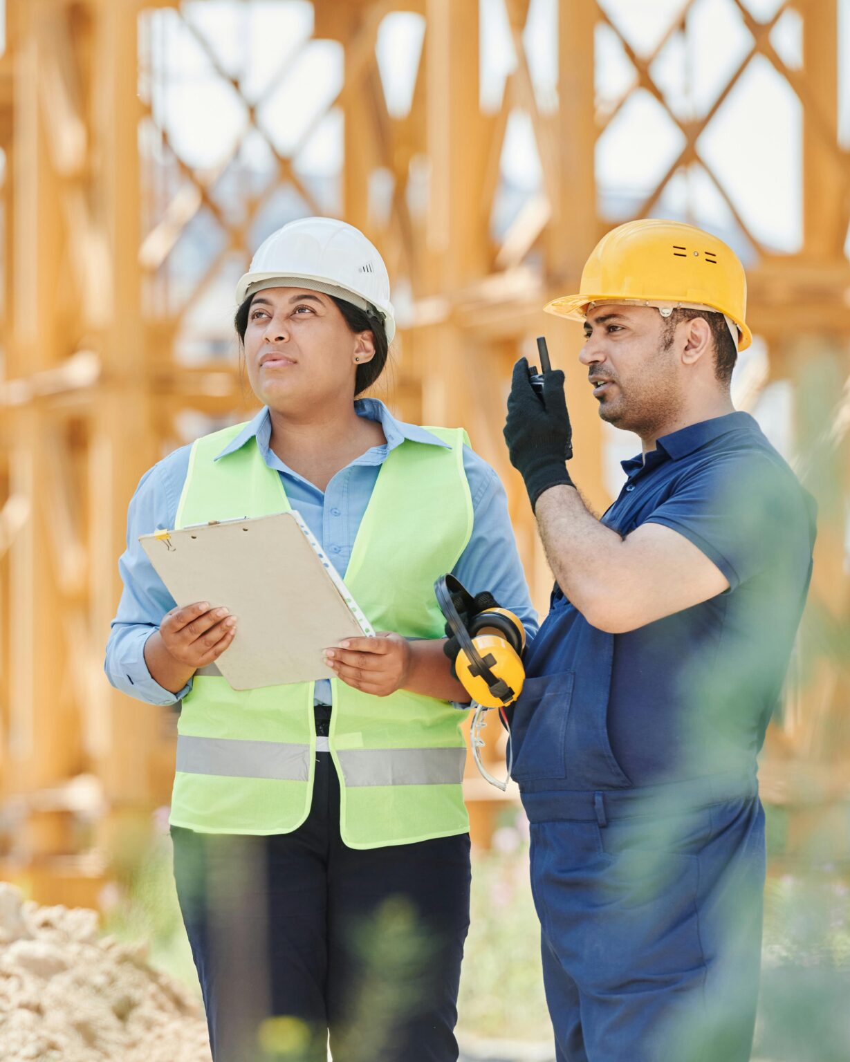 Image of two people, man and woman, surveying a jobsite. The woman is holding a clipboard while the man is speaking into a hand-held short-wave walki-talkie.